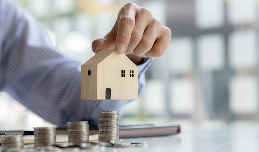 A hand holding a small wooden house over a stack of coins