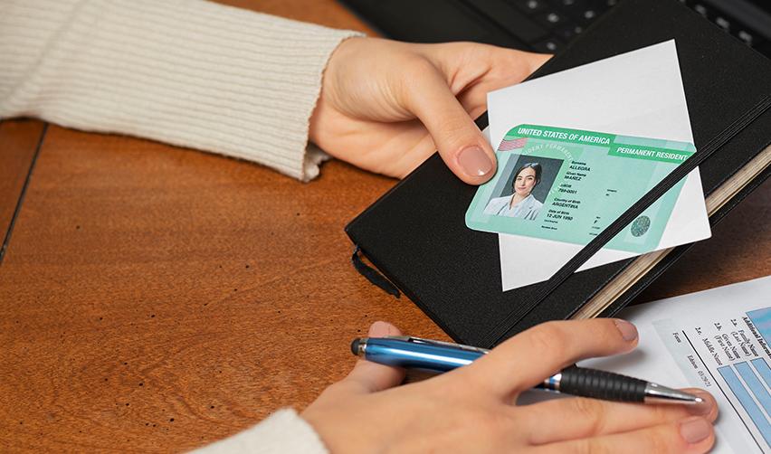 A person holding a pen and a Green Card, filling out an application.