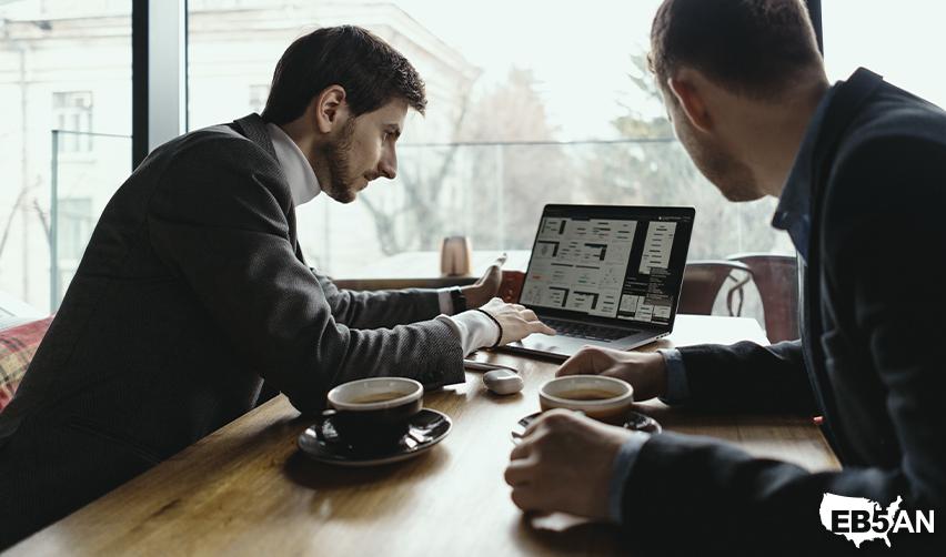 Two business professionals sitting at a table, analyzing investment data on a laptop while discussing EB5 visa opportunities and immigration strategies, with the EB5AN logo on the corner.
