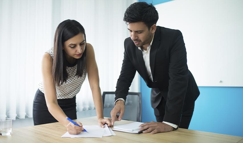 An Indian man in a suit reviewing documents with a female colleague in a corporate setting, illustrating the employment restrictions and sponsorship requirements of the H1B visa.