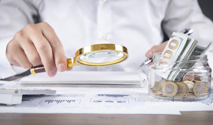 Close-up of an EB5 investor reviewing source of funds with magnifying glass beside a jar of cash and coins, symbolizing financial transparency and lawful investment documentation.
