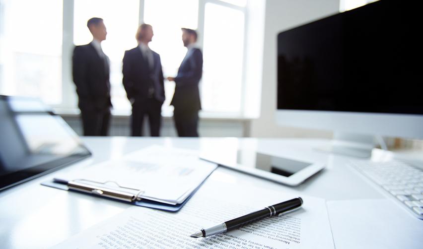 A legal office setup with documents and a pen in focus, while three professionals discuss strategy in the background—representing collaboration in EB5 legal planning.