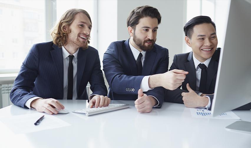 Three smiling professionals in suits reviewing immigration strategy data on a computer screen, representing collaborative EB5 investment planning.