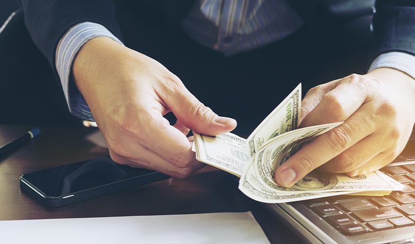 A businessman counting U.S. dollar bills at a desk with a phone and laptop, symbolizing the capital investment required for EB5 eligibility.