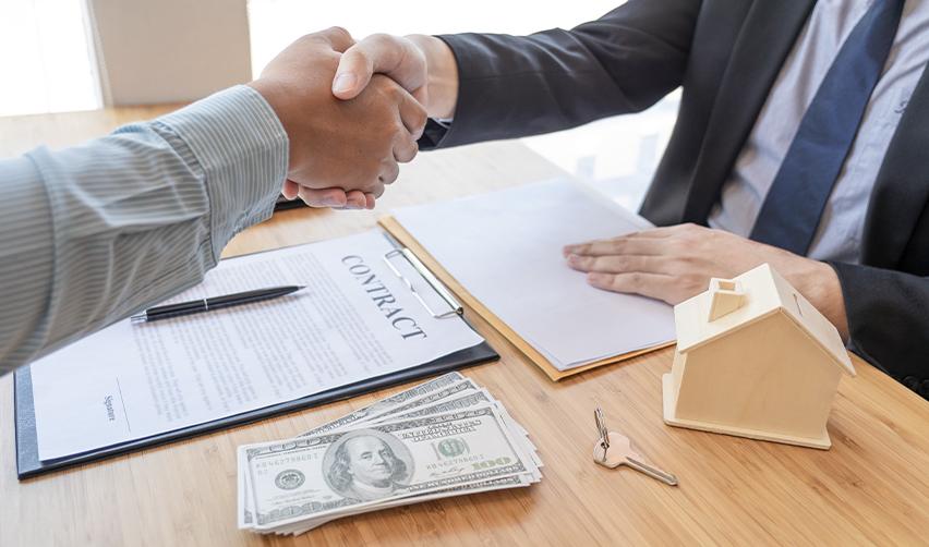 Close-up of two people shaking hands over a contract, cash, and a model house on a desk, representing EB5 investors using loans as part of a lawful source of funds.