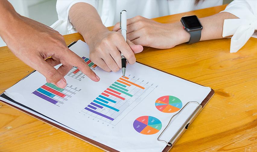 Hands pointing at bar charts and pie charts on a report, representing monthly EB5 project updates on construction, sales, and job creation.