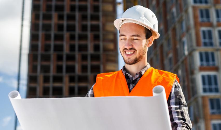 A construction engineer in a hard hat and orange vest reviewing project blueprints at a high-rise building site, symbolizing EB5 developers’ role in planning and execution.