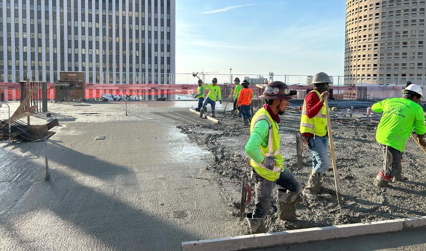 Construction crews leveling freshly poured concrete on an upper-level deck of ONE Tampa, with downtown buildings in the background showing active vertical progress on the EB5 project.